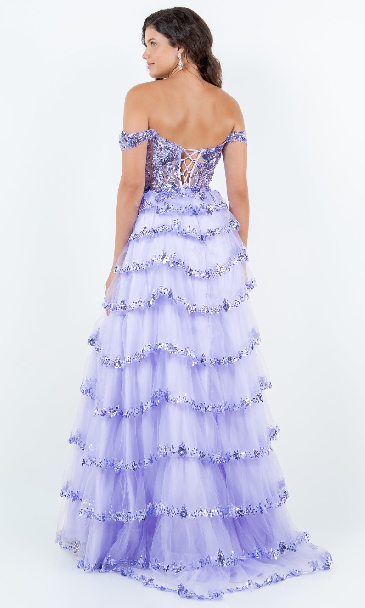 A woman faces away in the Ruffle Prom Ball Gown: Cinderella Couture 8228J, a lavender off-the-shoulder dress featuring a lace-up back, tiered ruffled skirt, sequin embroidery, and floral embellishments.